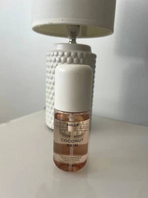 Phlur Coconut Skin mist - Fräsch parfym från Phlur med doft av kokos, frangipani och sandelträ. Flaskan är cylinderformad i genomskinligt glas med vit kork och ljusrosa vätska. Perfekt för dig som gillar tropiska och fruktiga dofter.