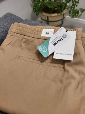 Beige mjukisbyxor från These Glory Days - Snygga beige mjukisbyxor från These Glory Days i mjukt och lätt material med elastisk midja och raka ben. Byxorna har sidofickor och en bakficka, perfekta för en chill och avslappnad stil. Tillverkade i Tencel för extra komfort.