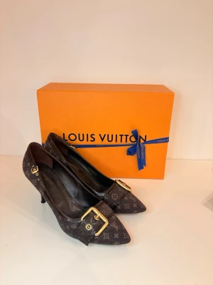 Bruna Louis Vuitton pumps med monogram - Säljer ett par bruna pumps från Louis Vuitton med klassiskt monogrammönster och spetsig tå. Skorna har en elegant guldfärgad spänndetalj framtill och smal klack. Materialet är tyg med läderdetaljer. Perfekta för dig som vill ha en lyxig och trendig look. Använd väldigt få gånger då jag ej är så bra på att gå i klackskor, säljer till ett väldigt bra pris då jag bara ej vill ha kvar dem, då jag inte kan gå i dem tyvär 🥲🥲