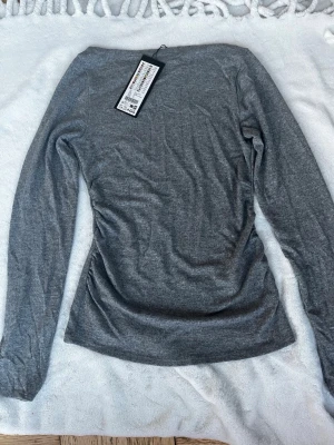 Grå långärmad topp - En enkel och stilren grå långärmad topp i mjukt material. Perfekt att matcha med jeans eller kjol för en clean look. Toppen går in vid midjan och har en boatneck. Helt ny och oanvänd, ny med prislapp kvar. Toppen är i storlek L men har stretchigt material som sitter tajt så funkar för personer med storlek S och m också