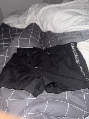 Svarta badbyxor från Calvin Klein - Snygga svarta badbyxor från Calvin Klein med logga längs sidan. Klassisk modell med dragsko i midjan och korta ben. Perfekta för stranden eller poolen. Materialet är snabbtorkande och lätt.