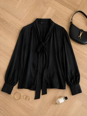 ZARA blus stl S  Tie-Neck - Ny oanvänd! A chic black satin blouse with a tie-neck detail and long puff sleeves, laid flat on a wooden floor. The blouse is styled with a black handbag with gold hardware, a pair of gold hoop earrings, a gold ring, and a small bottle of perfume.