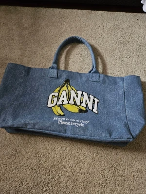 Blå shoppingväska med bananmotiv Ganni - Säljer en blå shoppingväska från Ganni i denimliknande material. Väskan har två handtag och ett stort bananmotiv med texten 'GANNI' samt 'Have a nice day! Please recycle' broderat på framsidan. Perfekt för dig som vill ha en unik och rymlig väska.