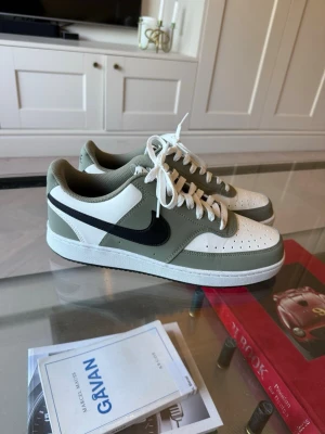 Nike Court Vision, oanvända  - Snygga sneakers i vitt och olivgrönt med svart swoosh på sidan. Skorna har vita snören, perforerad tåbox och en klassisk låg siluett. Yttermaterialet verkar vara en mix av läder och syntet. Perfekta för dig som gillar stilrena och sportiga vibes.