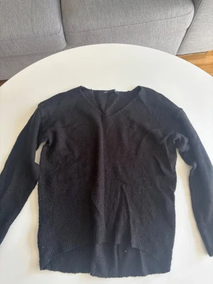 Svart stickad tröja med V-ringning - En enkel svart stickad tröja med V-ringning och långa ärmar. Tröjan har ribbade muddar vid ärmslut och nederkant samt något längre bakstycke. Perfekt basplagg till garderoben.