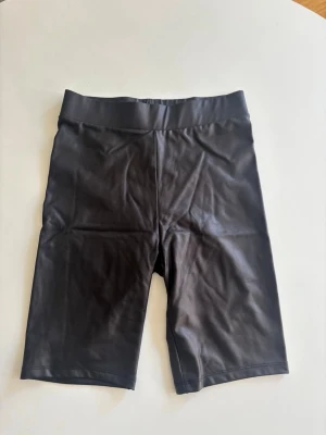 Svarta cykelshorts i stretchigt material - Säljer ett par svarta cykelshorts med hög midja och tight passform. Shortsen är gjorda i ett stretchigt, glansigt material som ger en sportig look. Perfekta för träning eller som en cool bas i garderoben.