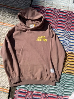 Brun hoodie från Gallery Dept. - Säljer en brun hoodie från Gallery Dept. med gul text på bröstet och en vit patch vid nederkanten. Hoodien har huva, magficka och ribbade muddar. Materialet är mjukt och skönt, perfekt för chill dagar. Oversized fit för en avslappnad stil.