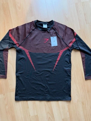 Onyx V5 - Snygg långärmad träningströja från Gymshark i svart med vinröda detaljer och mönstrade partier över axlar och armar. Tillverkad i ett stretchigt funktionsmaterial som andas, perfekt för gymmet eller löpning. Tight passform och sportig design.