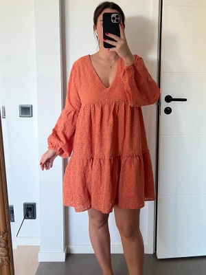 Klänning - Söt orange kortklänning med v-ringning och volangdetaljer. Klänningen har långärmade puffärmar och är tillverkad i ett lätt, perforerat tyg som ger en luftig känsla. Perfekt för varma dagar och sommarens alla tillfällen.