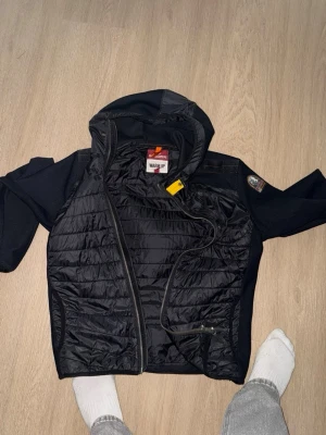 Parajumper cardigan storlek m men passar även s - Snygg svart pufferjacka med huva och quiltad front. Jackan har dragkedja, sidofickor med dragkedja och ett patch på ena ärmen. Materialet är en mix av glansigt tyg framtill och stickade ärmar och sidor. Perfekt för kalla dagar.
