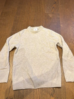 Beige stickad tröja i ullmix - Klassisk beige stickad tröja med rund hals och ribbade muddar vid ärmslut och nederkant. Tröjan har en enkel och stilren design, perfekt för lager-på-lager och passar till det mesta. Materialet ser ut att vara en mjuk ullmix. Den är lite mindre i storlek, skulle säga att 170-173 är rätt längd för tröjan!