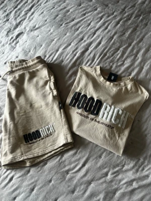 Beige set från Hoodrich med tryck - Säljer ett beige set från Hoodrich med matchande t-shirt och shorts. Båda plaggen har tydligt svart och vitt HOODRICH-tryck framtill och texten 'Product of the Streets'. T-shirten har rund hals och korta ärmar, shortsen har dragsko i midjan och sidofickor. Perfekt för en avslappnad streetwear-look. byxor= M t-shirt= S 