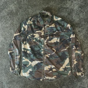 Camo overshirt från Replay - Snygg overshirt från Replay i camomönster med stjärnor. Skjortan har två bröstfickor med lock, krage och dragkedja framtill. Regular fit och tillverkad i ett lätt bomullstyg. Perfekt för lager på lager och streetwear-stil.