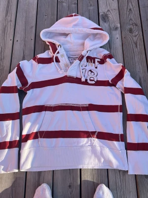 Randig hoodie från Abercrombie & Fitch - Säljer en vit hoodie med breda röda ränder från Abercrombie & Fitch. Tröjan har huva med snören, knappar vid halsen och en stor magficka. På bröstet står det 'ANF 18' i broderade bokstäver och siffror. Perfekt för en avslappnad och sportig stil.