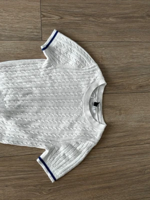 Vit kabelstickad t-shirt med blå kant - Vit kabelstickad t-shirt med korta ärmar och ribbade muddar. Ärmsluten och nederkanten har en blå rand som ger en sportig touch. Tröjan har rund halsringning och är gjord i mjukt stickat material.