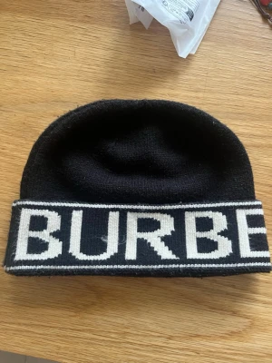 Svart mössa med Burberry-logga - Snygg svart mössa från Burberry med bred uppvikt kant och stor vit logga runtom. Tillverkad i mjukt stickat material som håller dig varm och ger en clean streetwear-look mössan går även att vända ut och in så backgrunden till texten blir vit.