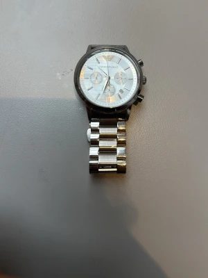 Emporio Armani silverfärgad klocka - Stilren klocka från Emporio Armani i rostfritt stål med silverfärgad länkarmband och vit urtavla. Klockan har kronograf-funktion, romerska siffror och tre mindre urtavlor. Perfekt accessoar för dig som gillar klassisk och elegant stil.