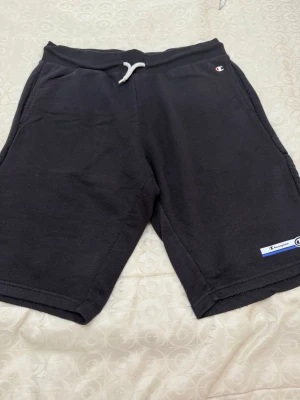 Svarta Champion shorts med dragsko - Ett par svarta shorts från Champion med vit dragsko i midjan och två sidofickor. Tillverkade i mjukt sweatshirtmaterial, perfekta för en avslappnad stil. Champion-logga finns både på benet och vid fickan.
