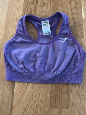 Lila sport-bh från Gymshark - Säljer en lila sport-bh från Gymshark med brottarrygg och ribbade detaljer. Materialet är stretchigt och andas, perfekt för träning. Diskret logga i vitt framtill. Snygg passform och bekväm resår under bysten.