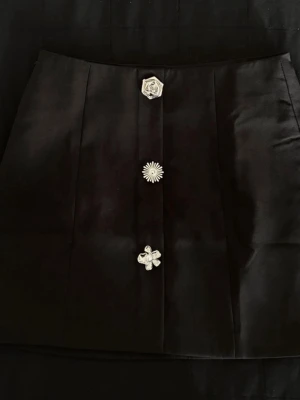 Elegant kjol med unik design - Säljer en svart elegant kjol med unik design, dekorerad med tre olika silverfärgade knappar framtill. Kjolen har en rak och kort form, perfekt för att skapa en cool och stilren look. 