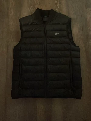 Svart dunväst från Lacoste - Snygg svart dunväst från Lacoste med klassisk ståkrage och dragkedja framtill. Västen har det ikoniska gröna krokodilmärket på bröstet och quiltad design som ger en sportig och modern look. Perfekt att slänga över en hoodie eller tröja för extra värme. Nypris 2000kr