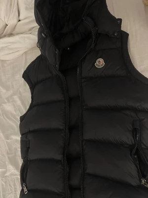 Svart dunväst med huva från Moncler - Snygg svart dunväst från Moncler med huva och två dragkedjeförsedda fickor. Västen har Moncler-logga på bröstet och quiltad design som ger en puffig look. Perfekt för lager-på-lager och håller dig varm under kyliga dagar.