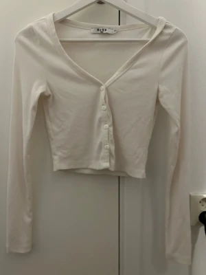 Vit ribbad cardigan från NA-KD crop top - Snygg vit ribbad cardigan från NA-KD med v-ringning och knappar framtill. Croppad modell med långa ärmar, perfekt att styla över ett linne eller t-shirt. Enkel och stilren design som passar till mycket.