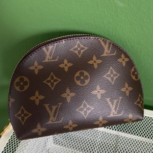 Brun trendig sminkväska el lite monogram necessär - Säljer en klassisk och trendig brun sminkväska eller liten necessär med ikoniskt monogrammönster i brunt Halvmåneformad design med dragkedja upptill. Tillverkad i tåligt material med lyxig känsla. Perfekt för smink eller småprylar.