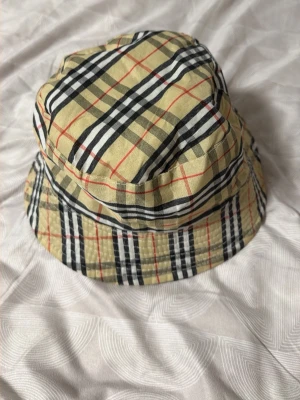 Buberry bucket hat i beige och svart - Säljer en trendig bucket hat med klassiskt rutigt mönster i beige, svart, vitt och röda detaljer. Hatten har mjuk form och är tillverkad i bomull, perfekt för att lyfta din outfit med en streetstyle-vibe.
