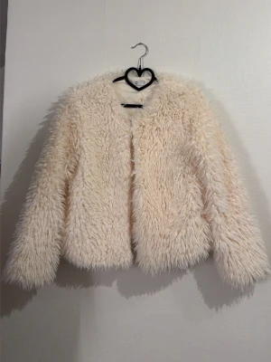 Krämvit fluffig pälsjacka - Säljer en supermjuk och fluffig pälsjacka i krämvit färg. Jackan har lång lugg, rund halsringning och raka ärmar. Perfekt för dig som vill ha en mysig och trendig look under vintern. Materialet är fuskpäls.