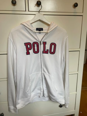 Vit hoodie från Polo Ralph Lauren - Vit hoodie med dragkedja från Polo Ralph Lauren. Stor röd POLO-text framtill, två fickor och huva. Klassisk och stilren design som passar perfekt till jeans eller joggers. Mjuk bomullskänsla och avslappnad passform.