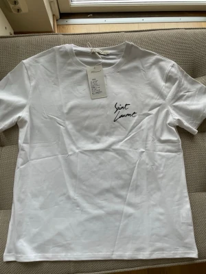 Vit Saint Laurent t-shirt - Snygg vit t-shirt med svart Saint Laurent-tryck både fram och bak. Klassisk rund hals och korta ärmar. Tillverkad i mjuk bomull som känns skön mot huden. Perfekt för en clean och stilren look.
