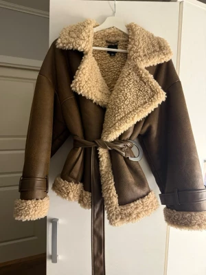 Brun teddy - Snygg brun kappa från Boohoo med mjukt teddyfoder i beige. Kappan har bred krage, bälte i midjan och detaljerade spännen vid ärmsluten. Använd endast en gång.