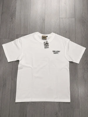 Vit Gallery Dept Tee - Det här är en klassisk Gallery Dept T-shirt som är helt ny med tags och påse. Allt följer med vid köp! Den är i storlek M och är true to size. Denna t-shirten är perfekt om du är ute efter streetwear stil eller bara för en vardags look, eller om man vill kan man ha den på specifika tillfällen. 