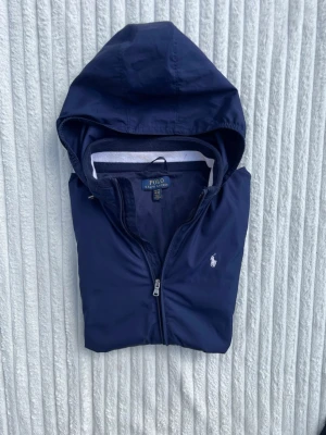 Marinblå vindjacka från Polo Ralph Lauren - Snygg marinblå vindjacka med huva från Polo Ralph Lauren. Jackan har dragkedja framtill, två fickor med zip och vit logga på bröstet. Elastiska muddar med vit detalj. Materialet är lätt och vattenavvisande, perfekt för blåsiga dagar.