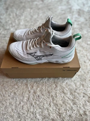 Vita Mizuno sneakers med gröna detaljer - Snygga vita sneakers från Mizuno med gröna detaljer på hälen och svart logga på sidan. Skorna har en sportig design med mesh- och syntetmaterial för bra andningsförmåga. Yttersulan har ett greppvänligt mönster, perfekta för träning eller handboll