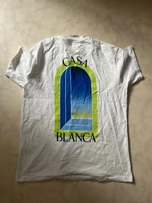  Casablanca T-shirt - Vit t-shirt från Casablanca med tryck av ett blått och gult fönster på både bröstet och ryggen samt texten 'CASA BLANCA'. T-shirten har rund hals och är tillverkad i mjuk bomull. Perfekt för en avslappnad och trendig stil.
