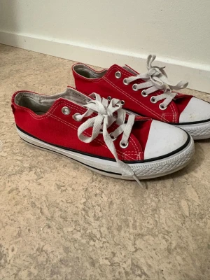 Röda Converse låga sneakers - Klassiska röda Converse sneakers i canvas med vita gummisulor och tåhätta. Skorna har vita snören och silverfärgade öljetter. Perfekta för en avslappnad och cool stil.