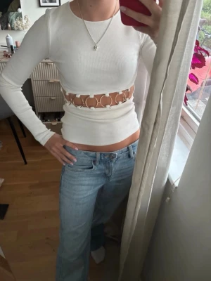 Vit långärmad topp med cut out-detalj - Trendig vit ribbad långärmad topp med rund halsringning och unika cut out-detaljer i midjan med ringar. Figurnära passform och stretchigt material som ger en snygg siluett. Perfekt för dig som vill sticka ut med en cool och modern look.