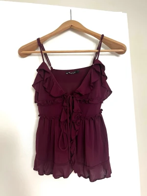 Vinröd volangtop från Gina Tricot - Supersöt vinröd topp från Gina Tricot med volanger, smala axelband och knyt framtill. Luftigt material och lätt transparent nedtill ger en romantisk vibe. Perfekt för dig som vill ha en feminin och trendig look. Endast använd 1 gång!
