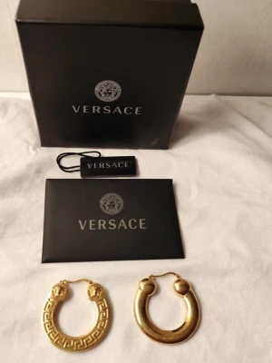 ELEGANTA TIDLÖSA GULDPLÄTERADE VERSACE ÖRHÄNGEN - En gåva jag aldrig hann ge bort:)  Guldpläterade Versace-örhängen med en Medusa-symbol och ett grekiskt nyckel mönster Greca-mönster.  Material: Örhängena är tillverkade av guldpläterad mässing.   * Örhängen är bly- och nickelfria och materialen är allergi vänliga.  VIKT: ca 17 gram per / örhänge.  Längd med lås: ca 3,7 cm  Bredd: 3,7 cm.  Tjocklek: ca 4 mm. Alla tillbehör som ses på bilden ingår, dock inge kvitto, därav priset.