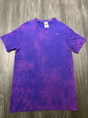 Gymshark Washed Seamless T-Shirt Purple, 2023 - Gymshark Washed Seamless T-Shirt – Purple, från AW23-kollektionen (2023). Snygg lila/rosa washed-design med figurnära muscle/slim fit-passform som framhäver fysiken. Sömlös konstruktion med ventilerande detaljer som ger hög komfort och rörelsefrihet under träning. Diskret Gymshark-logga på bröstet och en unik melerad finish. Perfekt för gym och träning.