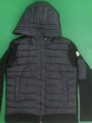 Svart pufferjacka med huva - Snygg svart pufferjacka med stickade ärmar och vadderad kropp. Jackan har en stor huva, dragkedja framtill och två fickor med knappar. Moncler-logga på vänster ärm. Perfekt för kalla vinterdagar och riktigt trendig look.