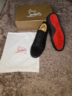Svarta Louboutin sneakers med nitar - Säljer ett par svarta sneakers från Christian Louboutin med klassisk röd sula och svarta nitar på tån. Skorna har lågt skaft, svarta skosnören och är tillverkade i mocka och läder. Perfekta för dig som vill sticka ut med en edgy och lyxig stil.