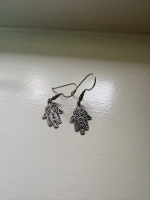 Silvriga örhängen med hamsa-händer - Unika örhängen i silverfärgad metall med detaljerade hamsa-händer. Hängande modell med fina mönster i handen, perfekta för dig som gillar boho eller spirituell stil. Lätta att bära och passar till många olika looks.