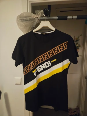 Svart Fendi t-shirt med logga och ränder - Säljer en svart t-shirt från Fendi med diagonal logga i brunt och vitt samt gula och vita ränder över bröstet. T-shirten har rund hals och korta ärmar. Perfekt för dig som gillar streetwear och vill sticka ut med en ikonisk design.