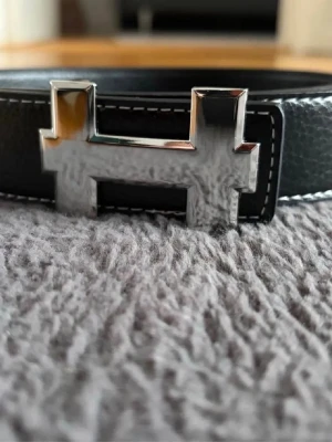 Svart bälte med silverfärgat H-spänne - Snyggt svart bälte med ett stort, blankt H-format spänne i silverfärg. Bältet har vit kontrastsöm längs kanterna och är gjort i ett slätt material som ger en stilren look. Perfekt accessoar för att lyfta din outfit.