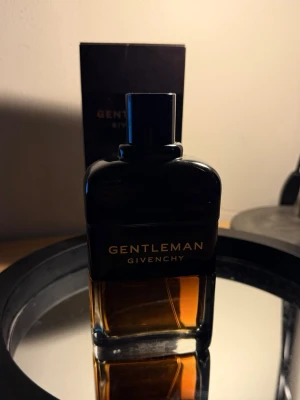 Givenchy Gentleman parfym - Givenchy Gentleman parfym i en elegant rektangulär flaska med svart ovandel och transparent nederdel som visar den bärnstensfärgade vätskan. Flaskan har stilren design med guldtext och svart lock. Perfekt för dig som gillar exklusiva dofter.