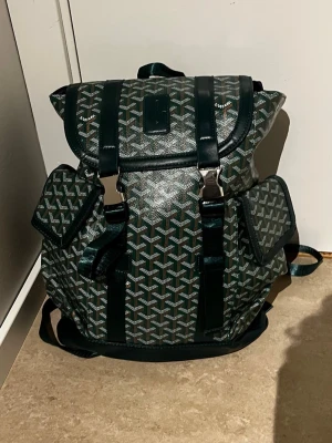 Mörkgrön gooyard mönstrad ryggsäck - Snygg mörkgrön ryggsäck med ikoniskt grafiskt mönster och detaljer i mörkgrönt läder. Ryggsäcken har två sidofickor med lock, justerbara axelband och metallspännen. Perfekt för dig som vill ha en unik och trendig accessoar till skolan eller stan.