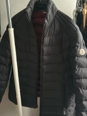 Svart dunjacka från Moncler - Snygg svart dunjacka från Moncler med quiltad design och dragkedja framtill. Jackan har två sidofickor med dragkedja och röd innerfoder med Moncler-logga. Perfekt för kalla vinterdagar och har en klassisk Moncler-patch på ärmen.